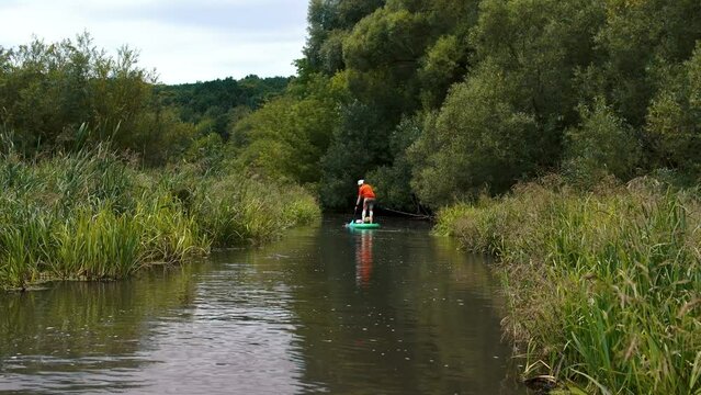 A narrow river among thickets of bushes.. Girl surfer in red t-shirt sitting on the sup board. Paddleboard SUP Water Sport Activity Holidays Vacation. Beautiful nature outdoors.