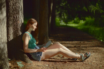 Side view of nice looking young woman in green top and blue shorts reading in the forest.
