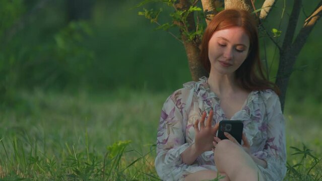 Redhead young girl using mobile phone apps sitting in green grass outdoors, makes purchases in online store. Ecommerce