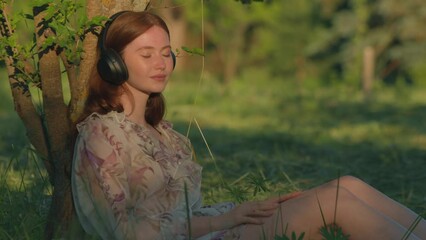 Girl in headphones listen to music sitting on green lawn in sunshine. Woman relaxing listening meditation outdoors