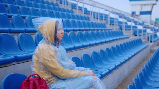 Young woman in raincoat sitting on stadium bleachers alone in rainy weather. Female spectator in wet waterproof coat at street stadium during competitions.