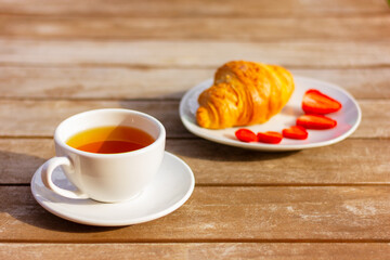 white cup with tea on saucer and plate with croissant and strawberry on wooden table in garden on sunny day, close up, selective focus