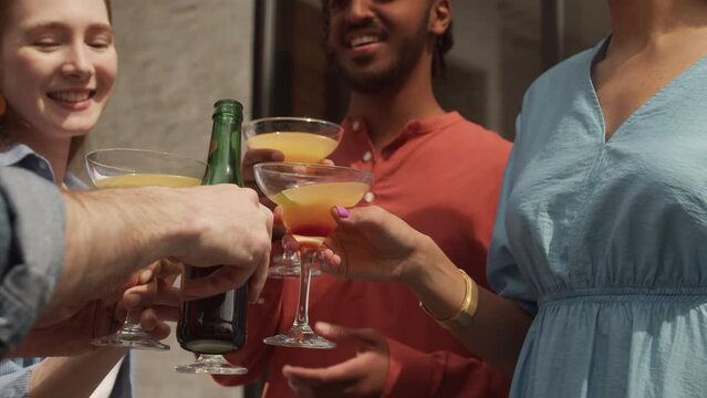 Slowmo closeup of multiethnic company of friends toasting with beer bottles and cocktail glasses while having outdoor terrace party on summer day