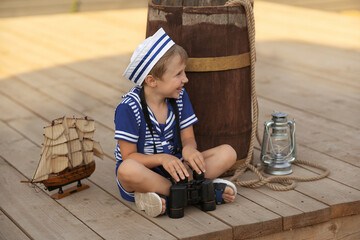 A boy of 7-8 years old in a sailor suit sits on a wooden deck against the background of a wooden...