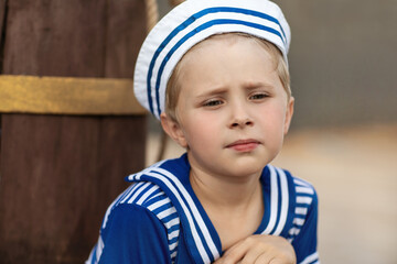 A close-up emotional portrait of a boy in a sailor suit and cap. The boy is sitting on the background of a brown wooden barrel. Summer, wood flooring. Emotions of a child