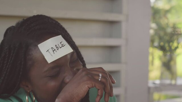 Medium close up with slowmo of young Black woman with word Table on sticker note on her forehead playing guessing game with friends outdoors in summertime
