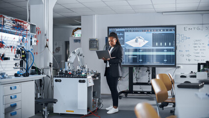 Black Female Automation Engineer Holding Laptop and Moving Bionic Arm With Written Code at Futuristic Factory. Modern Equipment and New Era in Computer Science and Robotics Industry Concept.