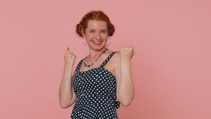 Happy redhead woman in polkadot dress shouting, raising fists in gesture I did it, celebrating success, winning, birthday, lottery goal achievemen. Young girl indoor on pink studio wall background