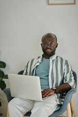 Portrait of African young businessman in eyeglasses sitting on armchair and working online on laptop