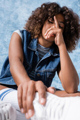 brunette african american woman in denim vest obscuring face with hand on blue background