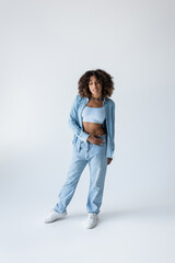 full length of african american woman in white sneakers and blue denim clothing on grey background