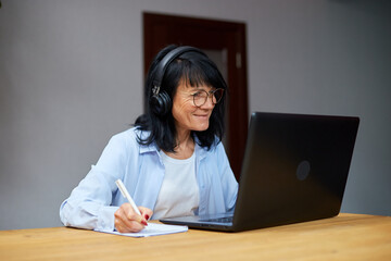 Smiling Mature woman wearing wireless headphones watch webinar