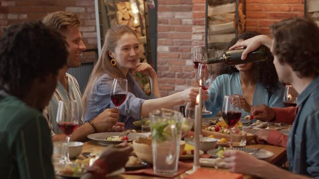 Medium slowmo of company of fancy multiethnic friends toasting with glasses of red wine while having festive dinner party in countryside house