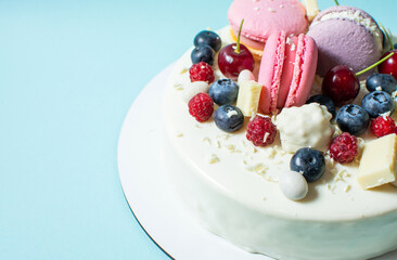 Tender white cake decorated with melted white chocolate, macaroons, berries and candies on blue background.