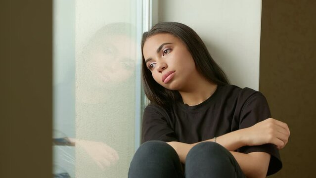 A sad black girl is sitting on the windowsill. A lonely young woman looks thoughtfully out the window. Concept of mental illness, problems.