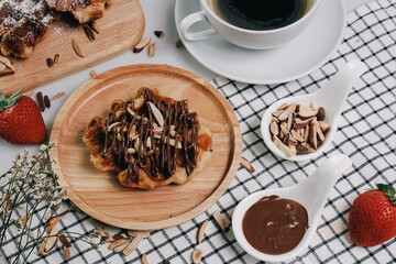 Croissant Waffle Korea pastry. Croffle with almonds and chocolate sauce topping served in wooden plate on square pattern table cloth. Selective focus.