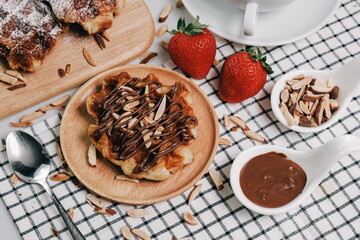 Croissant Waffle Korea pastry. Croffle with almonds and chocolate sauce topping served in wooden plate on square pattern table cloth. Selective focus.