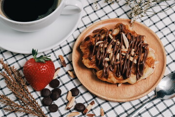 Croissant Waffle Korea pastry. Croffle with almonds and chocolate sauce topping served in wooden plate on square pattern table cloth. Selective focus.