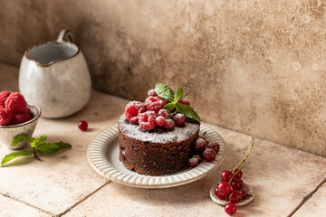 Chocolate cake with red summer berries, mint leaves and powdered sugar on beige tile background. Cozy still life, text space
