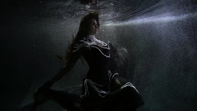 a long-haired woman in black clothes and pearl beads moves hands beautifully in dark water in bubbles. general plan