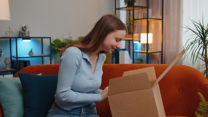 Happy young woman shopaholic unpacking delivery parcel sitting at home. Smiling satisfied pretty girl shopper online shop customer opening cardboard box receiving purchase gift by fast postal shipping