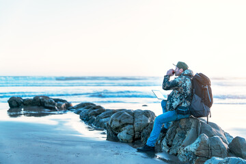 digital nomad sitting on the rock of the beach working with his laptop and drinking coffee relaxed.