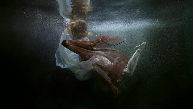 a red-haired woman in translucent fabrics floats and moves arms and legs beautifully under the surface of dark water. the general plan