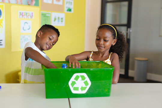 African american elementary students putting bottles in recycling container at desk in school