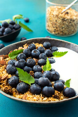 Homemade granola with yogurt, blueberries and mint leaves in a bowl on a turquoise background, delicious healthy breakfast