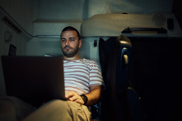 Young truck driver using laptop while resting in cabin at night.