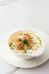 Norwegian red salmon fish soup with vegetables, herbs and cream, bowl of soup on a white marble background