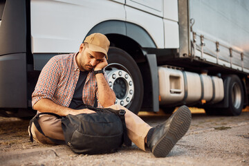 Displeased truck driver sitting on ground in front of his vehicle.