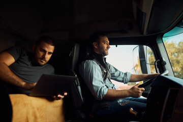Professional driver driving truck while his colleague is using laptop in cabin.