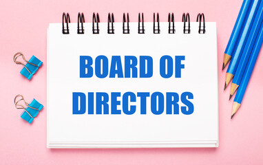 On a light pink background, light blue pencils, paper clips and a white notebook with the text BOARD OF DIRECTORS