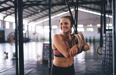 Fototapeta na wymiar Cheerful female bodybuilder with cute smile on face laughing during workout break near gymnastic rings, happy athlete keeping healthy lifestyle - spending weekend leisure in sportive gym studio