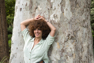 Portrait of an attractive mature woman with curly brown hair and shirt, leaning against a tree trunk with her hands on her head, seductive. Concept provoke, seduce, hairstyle, curls.
