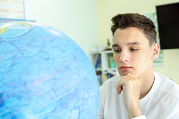 Portrait of a thoughtful young man in a white shirt sitting at a table in front of a globe. The concept of geography lesson, travel. High quality photo © Катерина Решетникова