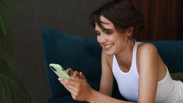 Young dreamful pensive minded woman wears casual clothes lying on blue sofa hold using mobile cell phone typing chat send sms stay at home rest relax spend free time in living room indoors grey wall