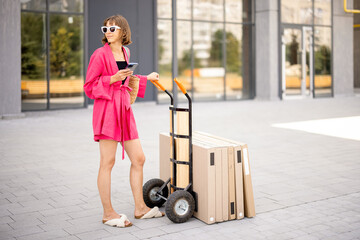 Woman using phone while standing near push cart with furniture in boxes, delivering them home....