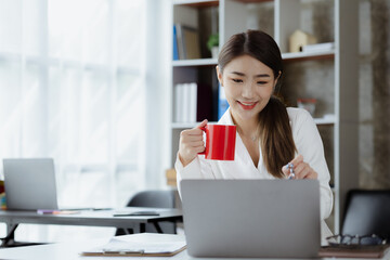 An Asian female employee sits with a red coffee mug in the startup's marketing department office, she is a marketing, customer liaison and consulting worker. Marketing concept.