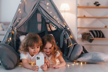 Using smartphone. Two little girls is in the tent in domestic room together