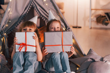 Holding gift boxes. Conception of presents and holidays. Two little girls is in the tent in domestic room together