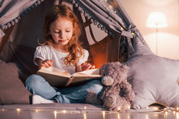 Reading the book. Cute little girl is in the tent in domestic room