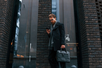 Holding smartphone. Stylish man with beard and in glasses is outdoors near building