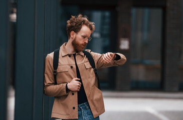 Looking at watches. Stylish man with beard in khaki colored jacket and in jeans is outdoors near...