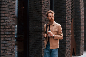 Coffee break. Stylish man with beard in khaki colored jacket and in jeans is outdoors near building