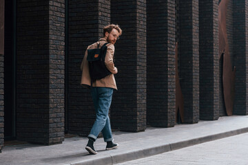 Stylish man with beard in khaki colored jacket and in jeans is outdoors near building