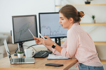 Using smartphone. Female stock broker in casual clothes is working in the office by pc