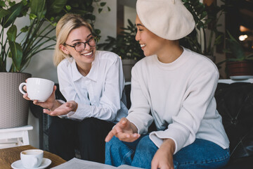 Joyful Asian and Caucaisan female friends dressed in stylish apparels discussing information and laughing during coffee meeting, cheerful hipster girls enjoying positive communication indoors