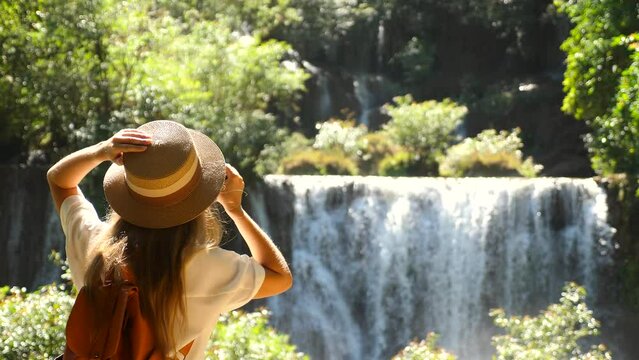 Female travel blogger with photo camera shooting amazing tropical waterfall in Asia journey. Cinematic and inspiration video of photographer woman taking picture of nature in travel vacation.